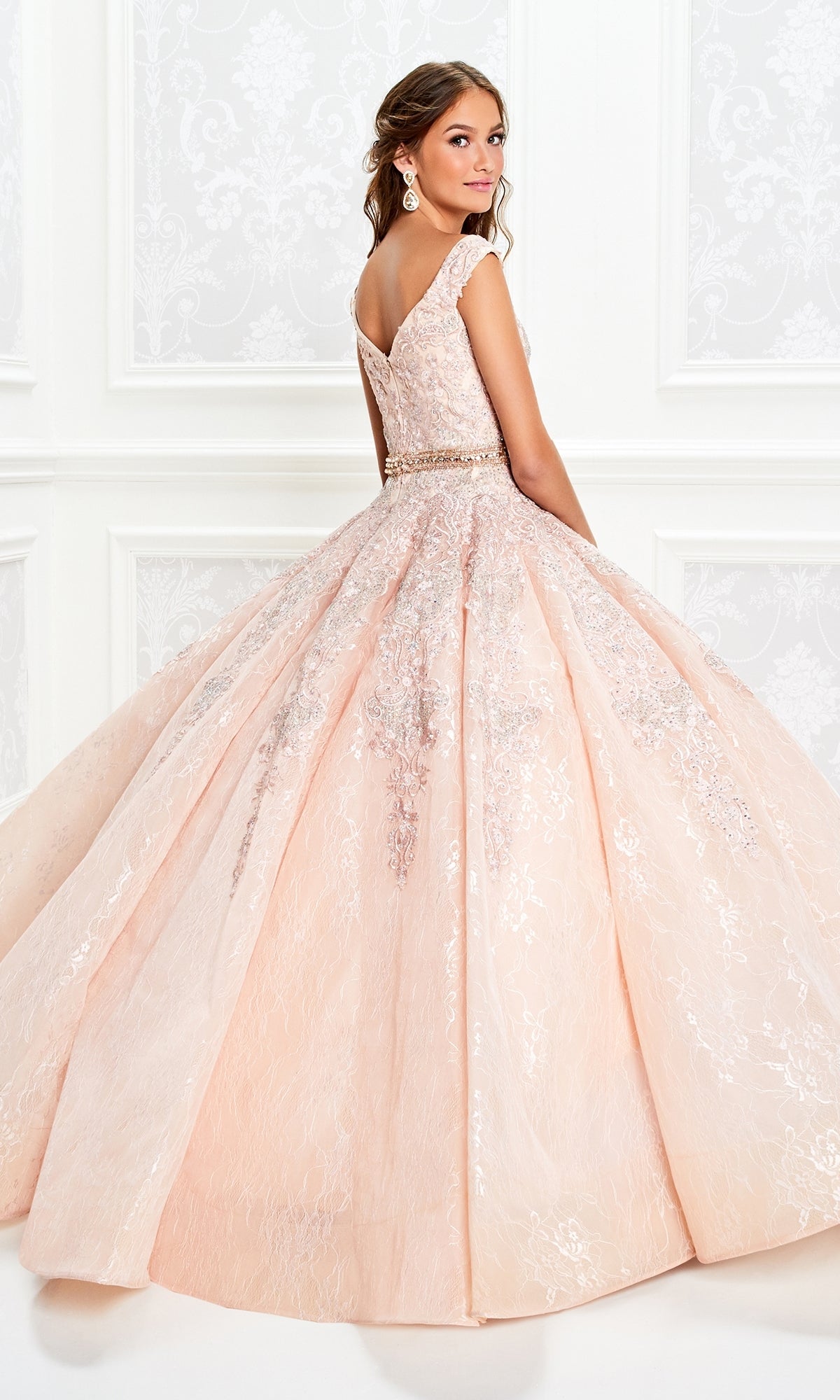 A young woman wears the Princesa Lace Ball Gown Quinceañera Dress PR11921, a light pink sparkly gown with a full skirt and beaded belt, as she stands in an elegant white room looking over her shoulder.